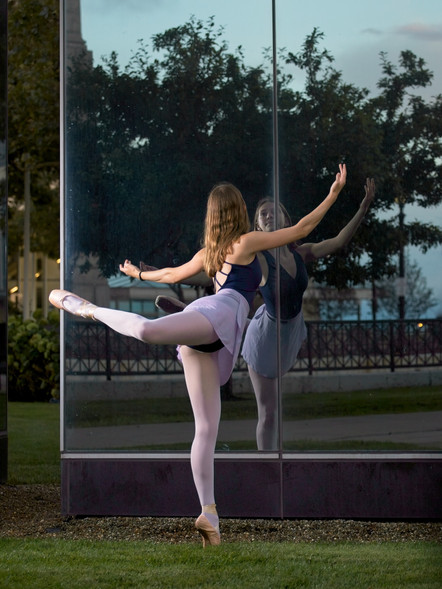 A ballet dancer performing an arabesque pose in front of a reflective glass wall, showcasing elegance and grace, with greenery in the background.