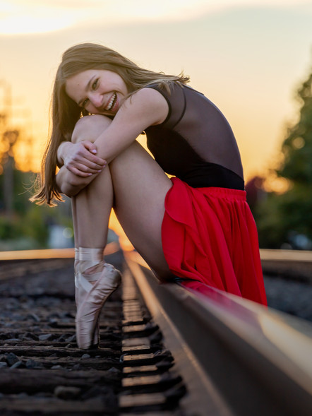 A young ballet dancer wearing a black top and a flowing red skirt sits on railway tracks at sunset, smiling and hugging her knees.