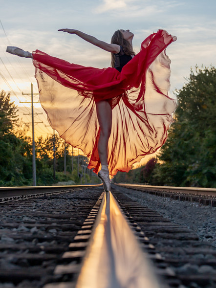 A female ballet dancer in a flowing red skirt leaps gracefully on a railway track during sunset, with vibrant colors in the sky and lush greenery in the background.