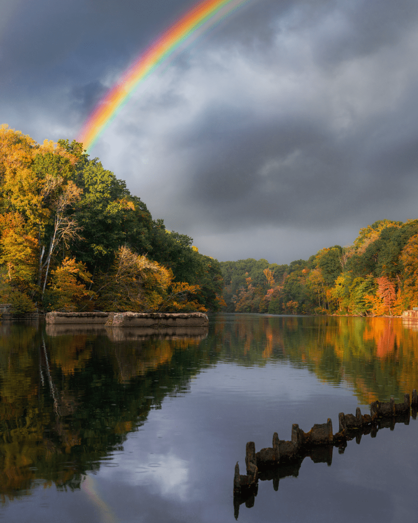 A serene landscape featuring a calm lake surrounded by vibrant autumn foliage, with a rainbow arching over the scene and gray clouds in the background.